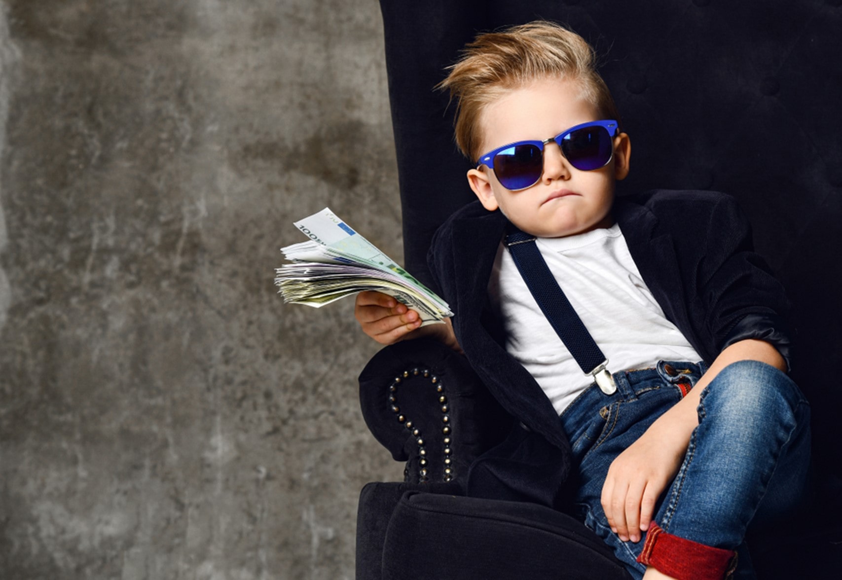 Young, cool, boy wearing sunglasses, leaning back in velvet chair, and holding a stack of cash.